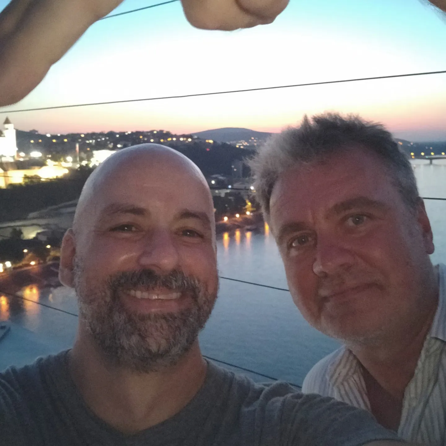 Two men smiling in front of a river at dusk, with a cityscape and a lit castle in the background. The atmosphere is relaxed and cheerful.